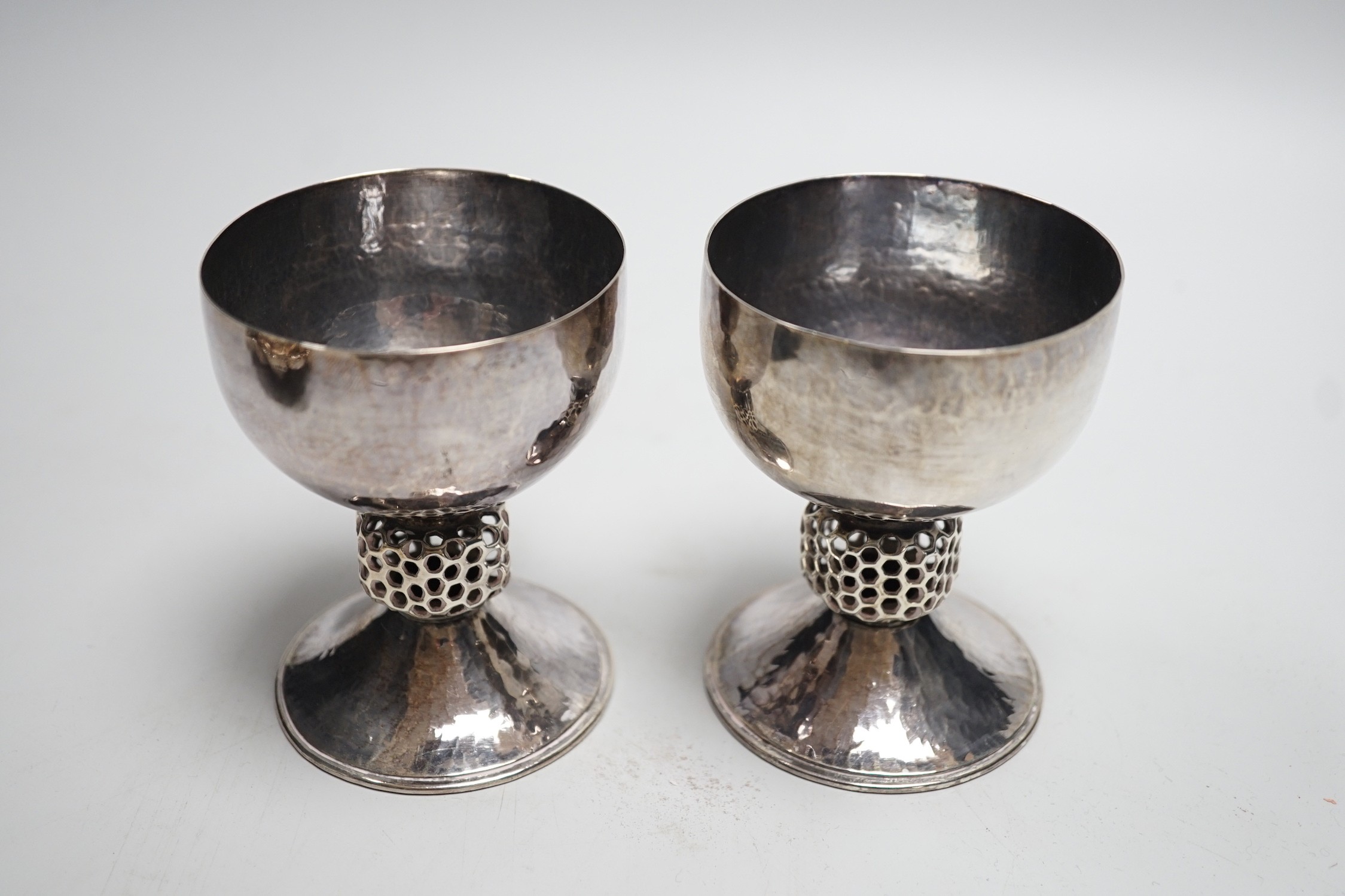 A stylish pair of Elizabeth II Arts & Crafts style planished silver pedestal cups, with reticulated stems, by Oliver Bailey, London, 1973, height 93mm, 7.5oz.